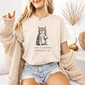 Puede incluir: Camiseta beige con una ilustración en blanco y negro de un gato sosteniendo una taza. El texto "I HAVE TO OVERTHINK THINGS OR I'LL DIE" está impreso debajo. Estilo casual, ideal para el día a día.
