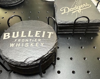 May include: A collection of slate coasters with white engraved text. One stack reads "BULLEIT FRONTIER WHISKEY", another says "Dodgers". The coasters are round and square, and are held in black metal holders.
