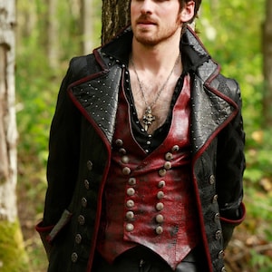 Puede incluir: Un abrigo negro y rojo de estilo steampunk con cuello alto y botones plateados ornamentados. El abrigo presenta un patrón acolchado en los hombros y ribetes rojos. Un chaleco rojo con botones plateados es visible debajo.