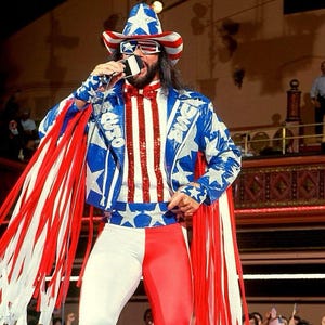 May include: A performer in a patriotic outfit, featuring a blue jacket with white stars, red and white fringing, and matching cowboy hat and sunglasses. The outfit includes red and white striped pants and a sequined top.