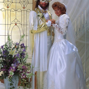 Puede incluir: Una pareja con atuendo de boda. La persona de la izquierda viste un traje blanco y dorado con sombrero y gafas de sol a juego. La persona de la derecha lleva un vestido de novia blanco con mangas de encaje y velo. Un arreglo floral está en primer plano.