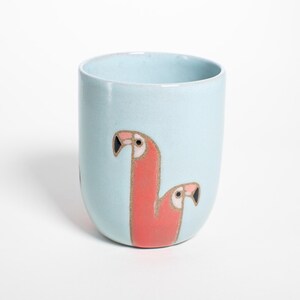 Op de afbeelding: Een lichtblauwe keramische mok met een afgeronde basis. De mok heeft een gestileerde illustratie van twee roze flamingo's met zwarte snavels. Het flamingo-ontwerp is goudkleurig omlijnd. De mok is effen van kleur.