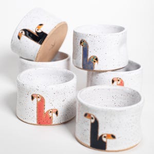 Puede incluir: Colección de tazas de cerámica hechas a mano con un acabado blanco moteado. Cada taza presenta un diseño de tucán único pintado a mano en varios colores, incluyendo negro, azul y coral. Las tazas son cilíndricas y tienen una estética rústica y artesanal.