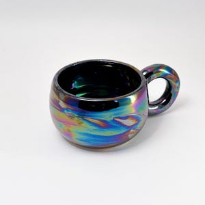 May include: Iridescent ceramic mug with a rounded body and a small handle. The mug's surface reflects a spectrum of colors, including blues, greens, and purples. The interior is dark, contrasting with the vibrant exterior. A unique and stylish drinkware item.