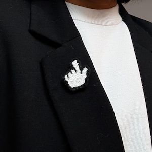 May include: A white and black embroidered patch shaped like a computer cursor hand icon, featuring a pointing finger design. The patch is attached to the lapel of a black blazer worn over a white ribbed shirt, creating a high-contrast visual.