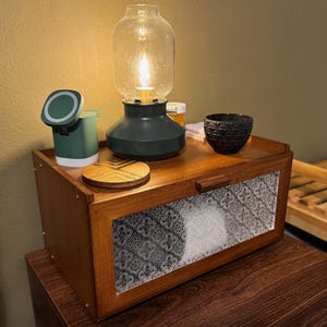 May include: A wooden nightstand with a decorative glass-topped lamp, a small green container, a wooden coaster, and a black textured bowl. The nightstand has a patterned glass panel on the front.