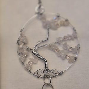 Può includere: Un ciondolo artigianale a forma di albero della vita in filo argentato. L'albero presenta un tronco e rami dettagliati, adornati con piccole pietre preziose di colore chiaro. Una sfera di cristallo sfaccettata è appesa sotto, sospesa da una catena d'argento.