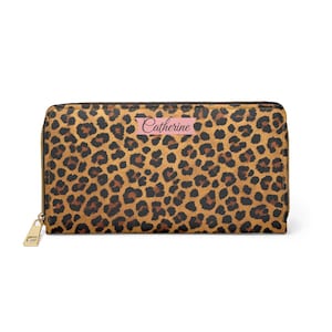 Puede incluir: Una cartera rectangular con estampado de leopardo con cremallera dorada y una etiqueta rosa que dice "Catherine". La cartera tiene un patrón de manchas marrones y negras.