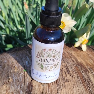 May include: A brown glass bottle with a black dropper, labeled "Hillybilly herbalist Dental Formula." The label features floral illustrations. The bottle sits on a wooden surface, with greenery and flowers in the background.