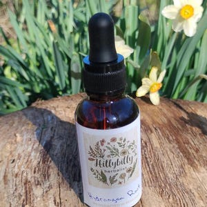 May include: A small, brown glass bottle with a black dropper top. The bottle has a white label with the text "Hillybilly herbalist" and "Hydrangea Root." The background features green foliage and yellow flowers.