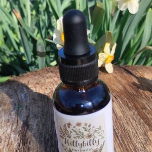 May include: A brown glass bottle with a black dropper, labeled "Hillybilly herbalist." The label features floral illustrations and text, including "Lavender + Mountain Mint Bitters." The bottle sits on a wooden surface, with daffodils in the background.