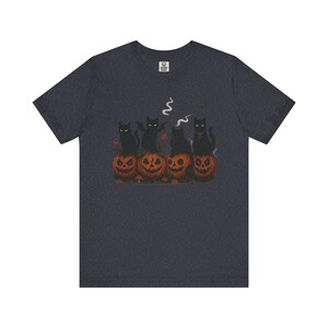 May include: Dark navy blue t-shirt featuring a Halloween design. Four black cats sit atop carved orange pumpkins. One cat has bat wings, and another has a collar. Wisps of smoke rise from the pumpkins, creating a spooky scene.