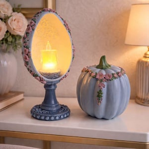 Può includere: Un portacandele decorativo a forma di uovo con una candela accesa all'interno e una zucca blu con accenti floreali. Il portacandele e la zucca sono su una superficie bianca, con una lampada e fiori sullo sfondo.