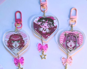 May include: Three heart-shaped keychains with cartoon character illustrations. Each keychain has a pink clasp, a pink bow, and a gold star charm. The keychains feature different characters with pink and purple color schemes.