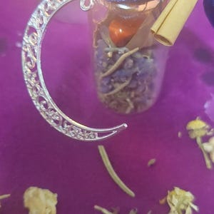 May include: A small glass bottle filled with dried herbs and a red stone, topped with a decorative lid and a silver crescent moon charm. A lit candle and purple surface are in the background.