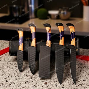 May include: A set of five kitchen knives with black blades and wooden handles. The handles have a unique design with a mix of light and dark wood, accented with a purple stripe. The knives are displayed in a black leather case.