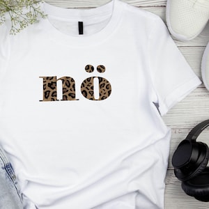Könnte beinhalten: Weißes T-Shirt mit dem Wort "nö" im Leopardenmuster. Das Shirt hat einen Rundhalsausschnitt und kurze Ärmel. Im Hintergrund sind weiße Turnschuhe und schwarze Kopfhörer zu sehen.