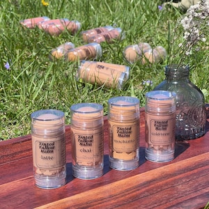May include: Four clear tubes of tinted tallow balm, labelled "latte", "chai", "machiatto", and "cold brew", are on a wooden tray. The tubes contain light brown balm. Other tubes are scattered in the grass in the background.