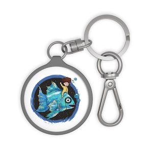 Könnte beinhalten: Ein runder Schlüsselanhänger mit einer Cartoon-Illustration einer Person, die auf einem blauen Fisch reitet. Das Design ist vor einem dunkelblauen Hintergrund mit Blasen angeordnet. Der Schlüsselanhänger hat einen silberfarbenen Ring und Verschluss.