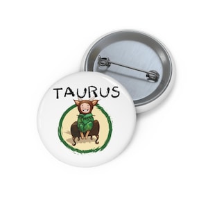 Puede incluir: Botón blanco con una ilustración de dibujos animados de una persona vestida de toro, con la palabra "TAURUS" en negro. La ilustración está dentro de un círculo verde. Un segundo botón es visible en el fondo.