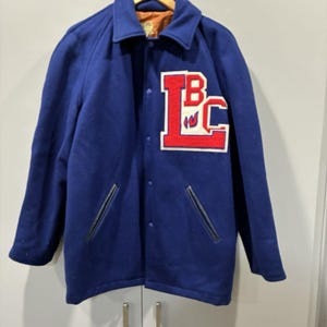 May include: A vintage blue wool letterman jacket with a red and white "BC" patch on the chest. The jacket has a button-up front, black leather-like accents on the pockets, and a classic collar.