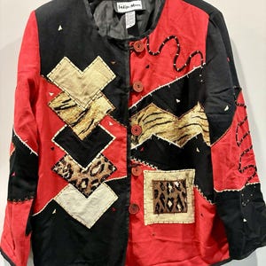 May include: A long-sleeve jacket by Indigo Moon featuring a color-blocked design in black and red. The front displays various square and chevron-shaped patches with beige, textured, and leopard print patterns, accented by decorative stitching and buttons.