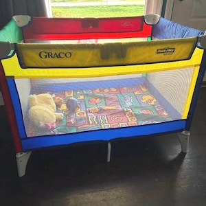 May include: A multi-colored Graco Pack 'n Play portable playard. The playard has a yellow, red, blue, and green frame with mesh sides. Inside, a colorful patterned mat and stuffed animals are visible.