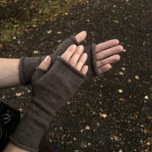 Könnte beinhalten: Ein Paar fingerlose Handschuhe aus braunem Strickstoff. Die Handschuhe weisen ein schlichtes glatt rechts gestricktes Muster mit gerollten Kanten an den Handgelenken und Fingeröffnungen auf und reichen bis zum Unterarm für Wärme und Schutz.