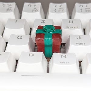 Könnte beinhalten: Ein farbenfroher, geschenkförmiger Keycap, rot und grün, liegt auf einer weißen Tastatur. Der Keycap ist ein Miniaturgeschenk mit einer grünen Schleife. Die Tastaturtasten sind weiß mit schwarzer Beschriftung.