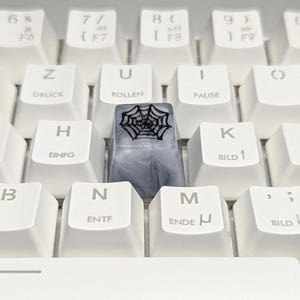 Könnte beinhalten: Nahaufnahme einer weißen Computertastatur mit einer individuellen Tastenkappe. Die Tastenkappe ist grau mit einem schwarzen Spinnennetz-Design. Die Tastaturtasten sind weiß mit schwarzer Beschriftung.