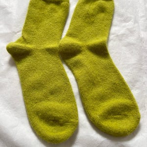 Puede incluir: Un par de calcetines verde lima. Los calcetines están hechos de un material suave y difuso. Los calcetines están doblados y colocados sobre una superficie blanca. Los calcetines son de color liso y parecen nuevos.