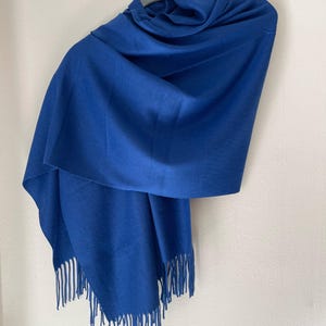 Puede incluir: Una bufanda azul vibrante con una textura suave y bordes con flecos, colgada de una percha gris. La bufanda está drapeada para mostrar su longitud y el detalle de los flecos en la parte inferior. Un accesorio sencillo y elegante.