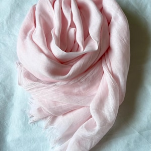 May include: A light pink scarf made of soft fabric, with a fringed edge. The scarf is draped in a circular pattern, creating a layered effect. The scarf is a solid color and appears to be lightweight.