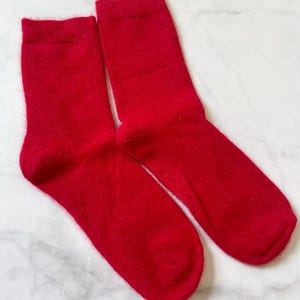 May include: A pair of bright red socks, made of a soft, fuzzy material. The socks are displayed on a white and gray marble surface. The socks are a solid color and appear to be ankle-length.