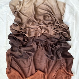 Puede incluir: Bufanda degradada marrón, que va del beige claro al chocolate oscuro y óxido. La tela es suave y fluida, con un borde con flecos. La bufanda se muestra sobre una superficie blanca, creando un contraste.