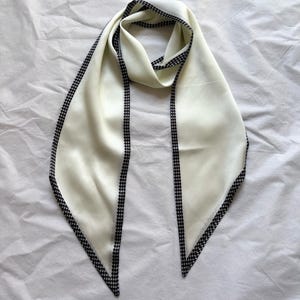 May include: Cream-coloured silk scarf with a black and white houndstooth trim. The scarf is displayed on a white surface, highlighting its smooth texture and elegant design. A stylish accessory.