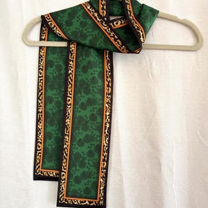May include: A long rectangular scarf featuring a deep green center with a black floral pattern. The scarf is bordered by a thin orange stripe and a wider band of black and tan leopard print, finished with a solid black edge trim.