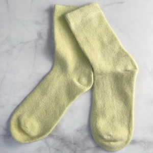 May include: A pair of pale yellow socks made from a soft, fluffy material. The socks are folded and rest on a white marble surface. The socks appear to be new and unworn.