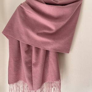 May include: A dusty rose scarf with a soft, textured appearance. The scarf is draped over a hanger, showcasing its fringed edges. The fabric appears to be a warm, cozy material, perfect for cooler weather.