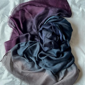 May include: A rectangular scarf featuring a gradient color transition from deep purple to dark blue and ending in a muted grey tone. The fabric has a soft, lightweight texture with delicate frayed edges along the perimeter of the entire piece.