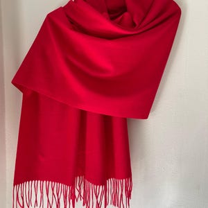 May include: A vibrant red scarf with a fringed edge, draped over a hanger. The scarf is made of a soft material and is a solid color. The scarf is a winter accessory.