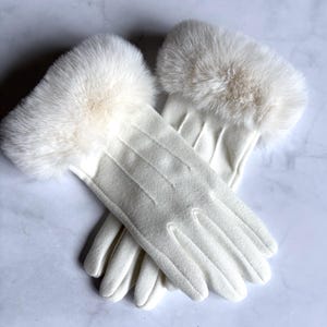 Peut inclure: Une paire de gants blanc cassé dotés d'un revers en fausse fourrure douce et moelleuse au poignet. Les gants sont fabriqués dans un tissu lisse de couleur unie avec trois lignes verticales cousues sur le dos de la main pour plus de détails.