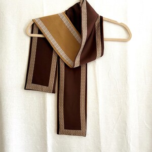 May include: A long rectangular scarf featuring a deep chocolate brown base colour with a contrasting tan section. The scarf is decorated with a repeating white geometric border pattern that frames the edges of both the brown and tan fabric segments.