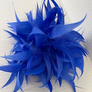 Pode incluir: Um acessório decorativo de penas azuis vibrantes. O item é composto por numerosas penas, criando uma forma esférica com bordas pontiagudas. As penas são de uma cor azul brilhante sólida, e a peça é provavelmente para uso decorativo.