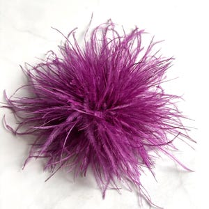 Può includere: Un accessorio in piume di struzzo color viola vibrante caratterizzato da una densa disposizione circolare di lunghe piume sottili che si irradiano verso l'esterno da un punto centrale, creando un aspetto pieno, strutturato e voluminoso.