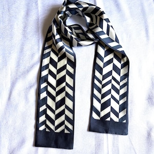 May include: A long, rectangular scarf featuring a repeating black and cream-coloured chevron pattern. The scarf has a solid black border at both ends and a smooth, satin-like finish, with the fabric draped to show the continuous geometric design.