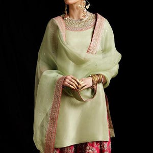 Può includere: Una tunica verde chiaro con scollo rotondo caratterizzata da ricami complessi, abbinata a un dupatta trasparente coordinato con bordo decorativo e pantaloni rosa vivace impreziositi da ricami floreali dettagliati e decorazioni metalliche.