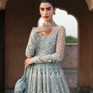 May include: A sage green floor-length gown featuring intricate silver embroidery, beadwork, and sequins. The dress has long sheer sleeves with floral patterns, a square neckline, and a matching drawstring pouch bag with silver embellishments.