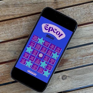 Op de afbeelding: Een smartphone met een "Epcot Bingo" spel op een blauwe achtergrond. Het spelbord heeft sterren en rechthoekige vakken. De telefoon ligt op een houten tafel met latten. De titel van het spel is wit.