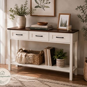 Op de afbeelding: Een witte sidetable met een donkerbruin houten blad, voorzien van drie lades met zwarte metalen handgrepen. De tafel heeft een onderste plank met een gevlochten mand met een crèmekleurige plaid, een stapel boeken en een kleine groene plant.